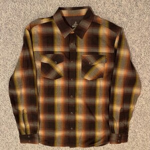 May include: A button-down, long-sleeve shirt in a plaid pattern of brown, orange, and yellow. The shirt has two chest pockets and a collar. The brand name "Prana" is visible on the collar.
