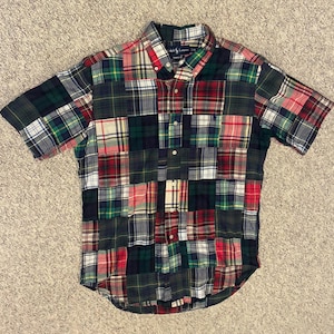 Può includere: Camicia patchwork a maniche corte con vari motivi a quadri. La camicia presenta un colletto button-down e una chiusura con bottoni. La tavolozza dei colori include tonalità di verde, rosso, blu e bianco. Realizzata in tessuto leggero.