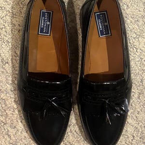 May include: A pair of black patent leather loafers with a glossy finish, featuring a kiltie fringe, a decorative tassel on the vamp, and a tan interior lining. The insole displays a blue and white label with the text Bostonian Classics.