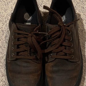 Könnte beinhalten: Ein Paar braune Lederschnürschuhe mit schwarzen Sohlen. Die Schuhe haben braune Schnürsenkel und einen gepolsterten Kragen. Die Schuhe sind für den Freizeitgebrauch und Outdoor-Aktivitäten konzipiert.