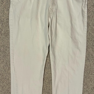 Puede incluir: Un par de pantalones vaqueros de color blanco roto con un diseño clásico de cinco bolsillos, cierre de botones y remaches metálicos en las esquinas de los bolsillos. Los pantalones tienen un corte recto con costuras visibles y cinturilla.