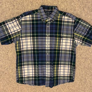 May include: A short-sleeved button-down shirt in a plaid pattern. The shirt features a mix of blue, green, white, and yellow squares. The shirt has a button-down collar and a small logo on the chest pocket.