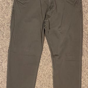 Puede incluir: Un pantalón verde oliva con un diseño clásico de cinco bolsillos. El pantalón tiene cierre de botón y cremallera, trabillas para el cinturón y bolsillos delanteros y traseros. La tela parece ser una mezcla de algodón duradera.