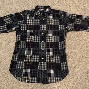 Può includere: Una camicia a maniche lunghe con bottoni e un design a patchwork. La camicia è realizzata con una varietà di tessuti a quadri in nero, bianco, grigio e marrone.