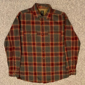 May include: A long-sleeved, button-down shirt in a plaid pattern of red, orange, and gray. The shirt has two chest pockets and a collar. The brand logo is visible on the collar. The shirt is made of a soft, possibly flannel, material.