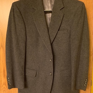 Peut inclure: Un blazer gris anthracite avec une fermeture à deux boutons et des revers crantés. La veste a deux poches avant et des boutons aux manches. Il est suspendu à un cintre en bois.