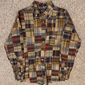 May include: A button-down shirt with a patchwork design. The shirt features a variety of plaid patterns in shades of blue, green, yellow, red, and brown. It has long sleeves, a button-down collar, and a chest pocket.