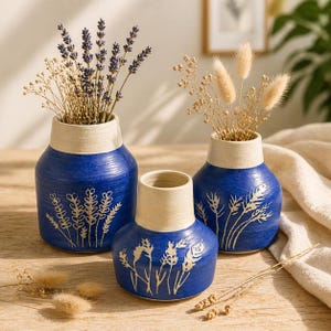 Könnte beinhalten: Drei Keramikvasen mit blauen Körpern und cremefarbenen Rändern. Jede Vase hat ein anderes botanisches Design in Creme. Die Vasen enthalten getrocknete Blumen und Gräser. Die Vasen sind auf einer Holzoberfläche angeordnet.