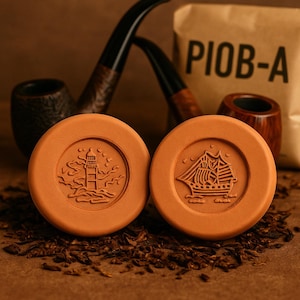 Peut inclure: Deux repose-pipes ronds, couleur terre cuite, avec des motifs en relief d'un phare et d'un voilier. Les supports reposent sur du tabac, avec deux pipes et un sac portant l'inscription "PIOB-A" en arrière-plan. L'image a une tonalité chaude et terreuse.