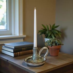 Könnte beinhalten: Ein Kerzenhalter aus Keramik mit einer weißen Spitzkerze. Der Halter ist in Grün- und Brauntönen glasiert und hat einen Griff. Der Kerzenhalter steht auf einer Holzoberfläche, im Hintergrund Bücher und Pflanzen.