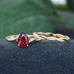 May include: A pear-shaped red gemstone engagement ring set with a twisted band and a matching wedding band. Both bands are set with small diamonds.