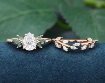 May include: Two rose gold rings. One ring features a pear-shaped, clear gemstone with green accents. The second ring has a band with small, clear gemstones arranged in a leaf-like pattern. The rings are displayed on a dark blue surface.