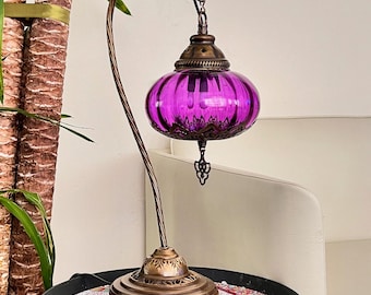 Op de afbeelding: Een paarse glazen tafellamp met een gebogen metalen arm en een sierlijke voet. De lampenkap heeft een textuur en een decoratief metalen knopje.