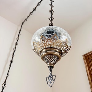 May include: A hanging pendant light with a globe-shaped, crackled glass shade. The shade is accented with a gold filigree pattern and a bronze-colored metal base. The light hangs from a black chain.