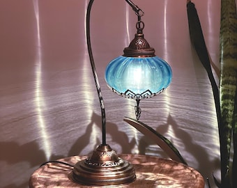 Könnte beinhalten: Eine blaue Glastischlampe mit einem gebogenen Arm und einem dekorativen Sockel. Die Lampe ist eingeschaltet und strahlt ein warmes Licht aus.
