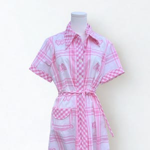 May include: A knee-length shirt dress in white with a pink plaid pattern and embroidered pink butterflies. The dress features short sleeves, a pointed collar, a button-down front, a matching fabric waist tie, and a single patch pocket on the front.
