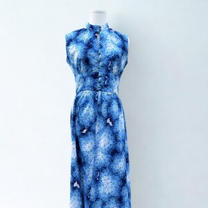 May include: A sleeveless floor-length dress featuring a vibrant blue and white floral pattern. The garment has a high collar with a small front slit, a fitted waist, and a vertical row of matching blue buttons extending from the neck to the waist.