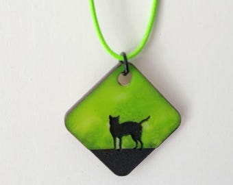 May include: A pendant necklace featuring a black cat silhouette on a green and black diamond-shaped pendant. The pendant is suspended from a bright green cord. The cat is standing on a black surface.