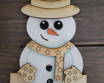 May include: Wooden snowman ornament with a brown hat, white body, orange nose, black eyes, and a scarf with white stars. The snowman is holding a star-shaped ornament in each hand. The ornament has the name "Elysia" written on the base.