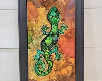 May include: A framed artwork featuring a green gecko with a black outline, set against a background of brown, orange, and yellow abstract ink washes.
