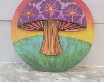 May include: A round wooden wall hanging with a painted image of a purple mushroom with a yellow and orange sunset background.