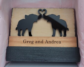May include: Wooden plaque with two black elephant silhouettes facing each other, their trunks forming a heart shape. The plaque has a smaller wooden plaque below it with the text "Greg and Andrea" engraved on it.
