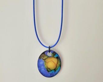 May include: A round pendant necklace with a blue cord. The pendant features a colorful design with shades of blue, yellow, and green. The pendant is approximately 1 inch in diameter.