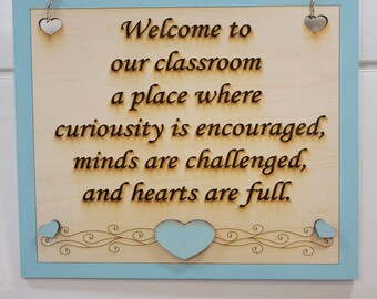 May include: Wooden sign with a light blue border and the text "Welcome to our classroom a place where curiosity is encouraged, minds are challenged, and hearts are full."