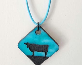 May include: A turquoise pendant necklace featuring a black cow silhouette on a diamond-shaped charm. The charm has a black border and a light blue cord. The cow is standing on a black base.