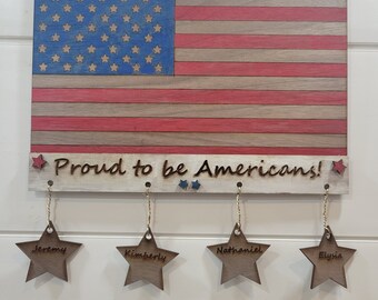 May include: A wooden American flag with red, white, and blue stars and stripes. The flag is hanging from a string and has a sign below it that says "Proud to be Americans!" with four wooden stars hanging below the sign, each with a name on it: Jeremy, Kimberly, Nathaniel, and Elysia.