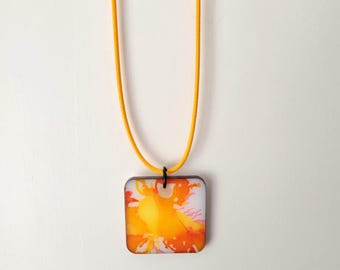 May include: Square pendant necklace with a vibrant abstract design in shades of orange and yellow. The pendant is suspended from a bright yellow cord. The necklace is displayed against a white background.
