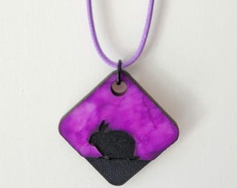 May include: A purple and black pendant necklace featuring a diamond-shaped charm. The charm has a purple watercolor background with a black silhouette of a rabbit. The necklace has a thin, lavender-colored cord.