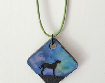 May include: A square pendant necklace featuring a black silhouette of a dog against a blue and purple watercolor background. The pendant is attached to a green cord. The dog is standing on a black base.