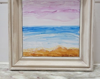 May include: A framed abstract painting depicting a seascape with a pink and white sky, blue water, and a sandy beach.