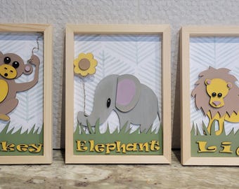 May include: Three wooden framed wall art pieces featuring a monkey, elephant, and lion. Each piece has a green grass background and the animal's name is written in yellow letters below the animal.