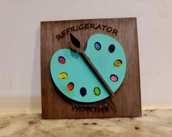 May include: A wooden sign with a painted artist's palette and paintbrush. The sign reads "Refrigerator Worthy".