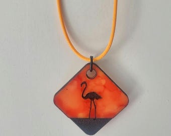 May include: A necklace featuring a pendant with a flamingo silhouette against an orange and black background. The pendant is diamond-shaped and hangs from a yellow cord.