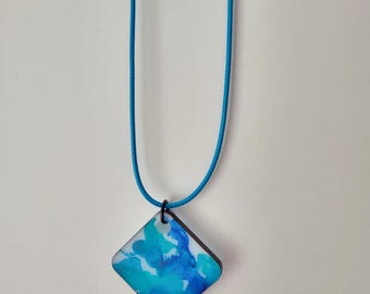 May include: A square pendant necklace with a blue and white watercolor design. The pendant is suspended from a turquoise cord. The pendant has a dark brown border and a black clasp.