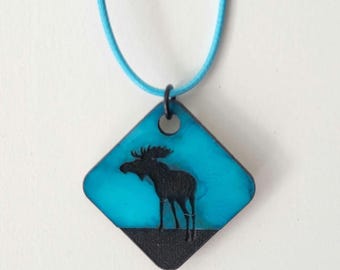 May include: A square pendant necklace featuring a black moose silhouette against a turquoise background. The pendant is attached to a light blue cord. The bottom of the pendant is black.