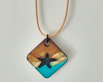May include: A diamond-shaped pendant necklace featuring a black starfish over a turquoise blue and gold-toned ocean scene. The top portion is a warm brown color. The pendant is suspended from a tan cord.