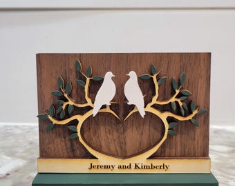 May include: A wooden plaque with a heart-shaped cutout and two white doves perched on a green branch. The plaque is on a green base and has the text "Jeremy and Kimberly" engraved on the bottom.