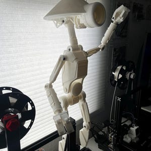 Könnte beinhalten: Ein weißes 3D-gedrucktes Modell eines Roboters mit einem großen Kopf und einer einzelnen Antenne. Der Roboter steht auf einer schwarzen 3D-Druckplattform.