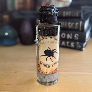 May include: A clear glass bottle labeled "Spider Eggs" with a black spider illustration. The bottle has a black dripped wax top and contains small, round, white objects. A decorative spider charm hangs from the bottle.