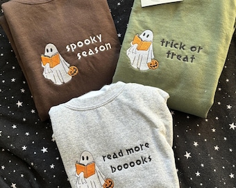 May include: Three sweatshirts with different Halloween-themed embroidery designs. The first sweatshirt is brown with a ghost holding a book and the text "spooky season". The second sweatshirt is green with a ghost holding a book and the text "trick or treat". The third sweatshirt is gray with a ghost holding a book and the text "read more booooks".