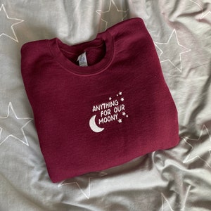 May include: A maroon sweatshirt with the phrase "ANYTHING FOR OUR MOONY" embroidered in white, featuring a crescent moon and stars design. The sweatshirt is folded on a gray star-patterned bedspread.