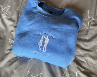 Op de afbeelding: Een lichtblauw sweatshirt met de tekst "slow dance when it rains" en een wit geborduurd beeld van twee dansende mensen.