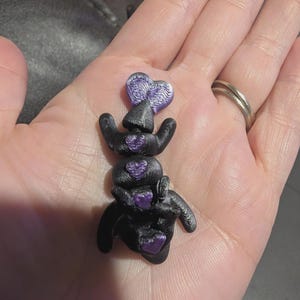 K&ouml;nnte beinhalten: Eine kleine, schwarze, bewegliche Figur mit strukturierter Oberfl&auml;che, die vier eingebettete, violette, herzf&ouml;rmige Akzente entlang ihrer Mitte aufweist. Das Objekt hat ein geschichtetes, 3D-gedrucktes Aussehen mit einer deutlich gerippten Textur.