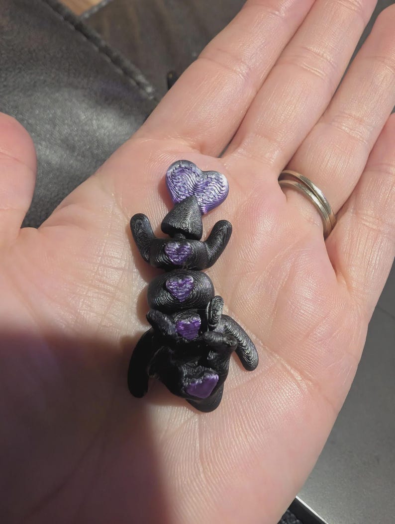 K&ouml;nnte beinhalten: Eine kleine, schwarze, bewegliche Figur mit strukturierter Oberfl&auml;che, die vier eingebettete, violette, herzf&ouml;rmige Akzente entlang ihrer Mitte aufweist. Das Objekt hat ein geschichtetes, 3D-gedrucktes Aussehen mit einer deutlich gerippten Textur.