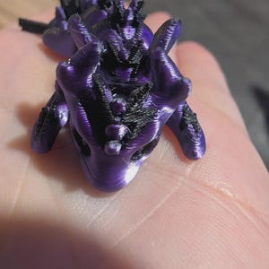 K&ouml;nnte beinhalten: Eine kleine, 3D-gedruckte Drachenfigur mit einem metallisch violetten K&ouml;rper und schwarzen Akzenten entlang des R&uuml;ckens, des Kopfes und der Gliedma&szlig;en. Das Wesen weist eine glatte, geschichtete Textur auf, die f&uuml;r die additive Fertigung charakteristisch ist.