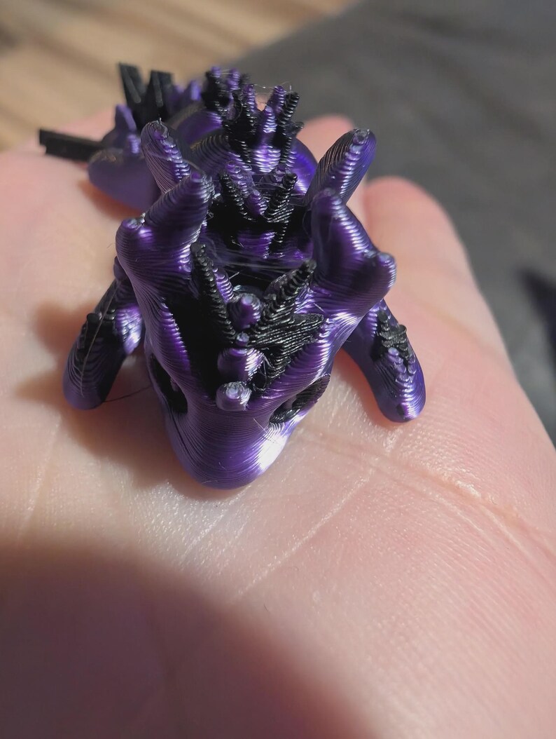 K&ouml;nnte beinhalten: Eine kleine, 3D-gedruckte Drachenfigur mit einem metallisch violetten K&ouml;rper und schwarzen Akzenten entlang des R&uuml;ckens, des Kopfes und der Gliedma&szlig;en. Das Wesen weist eine glatte, geschichtete Textur auf, die f&uuml;r die additive Fertigung charakteristisch ist.
